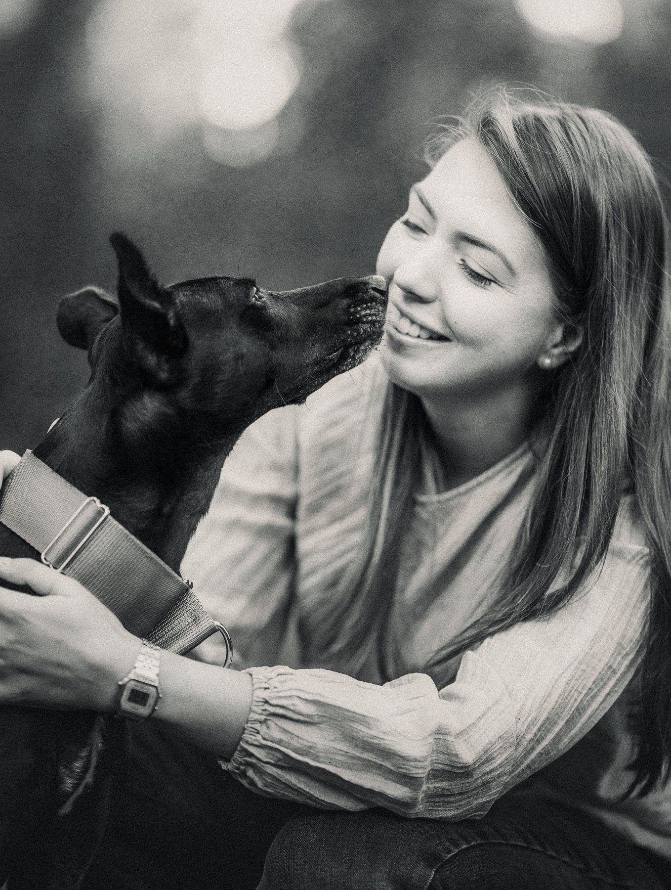 Zwart-witfoto van een glimlachende vrouw die van haar zwarte hond een kusje op haar wang krijgt.