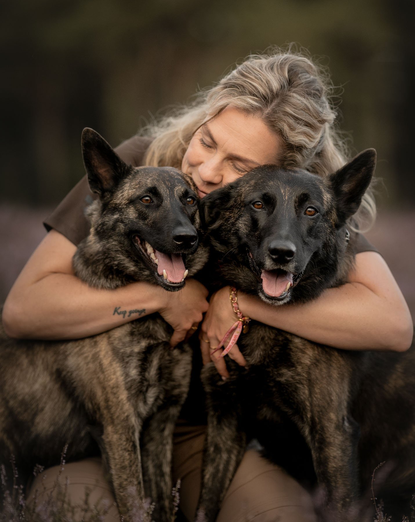 A woman with long grey hair hugging two dogs. The dogs have black and tan coats and look happy. The woman has a tattoo on her forearm and wears a bracelet.