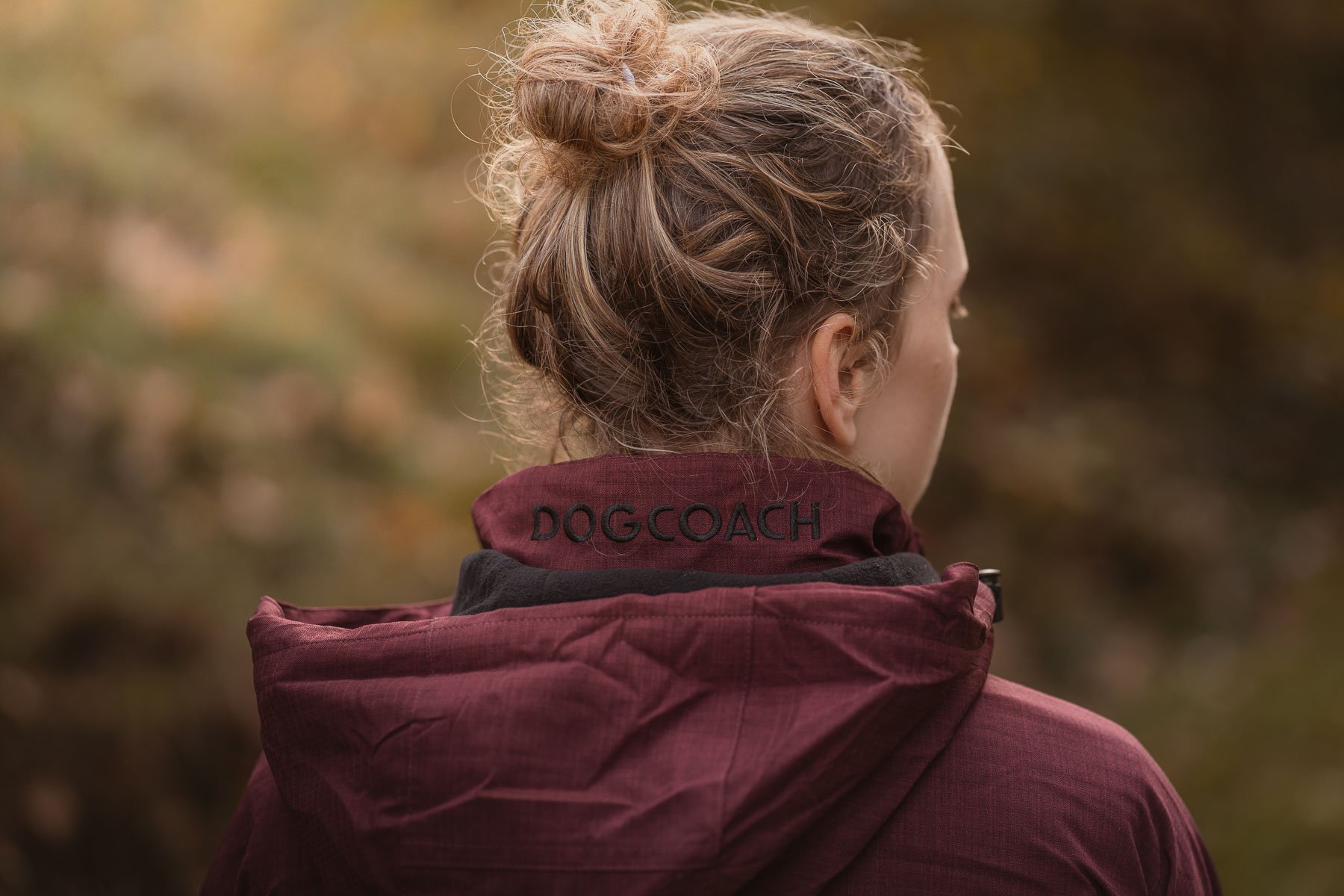 A woman looks away, wearing a red jacket with the text “DOGCOACH” on the collar, in a wooded setting with autumn leaves.