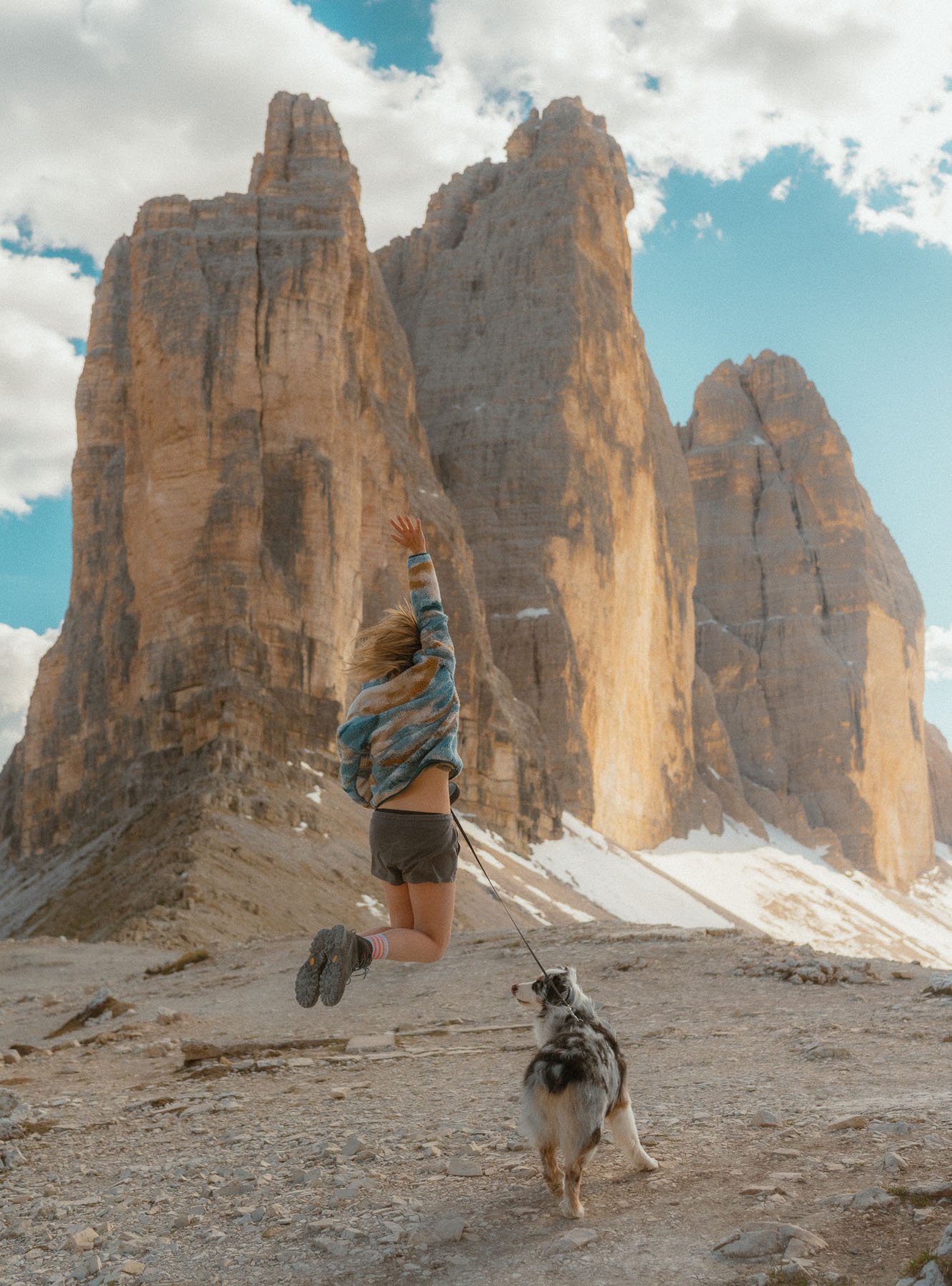 A person with a dog jumps into the air in a mountainous setting with rocks and snow, under a partly cloudy sky.