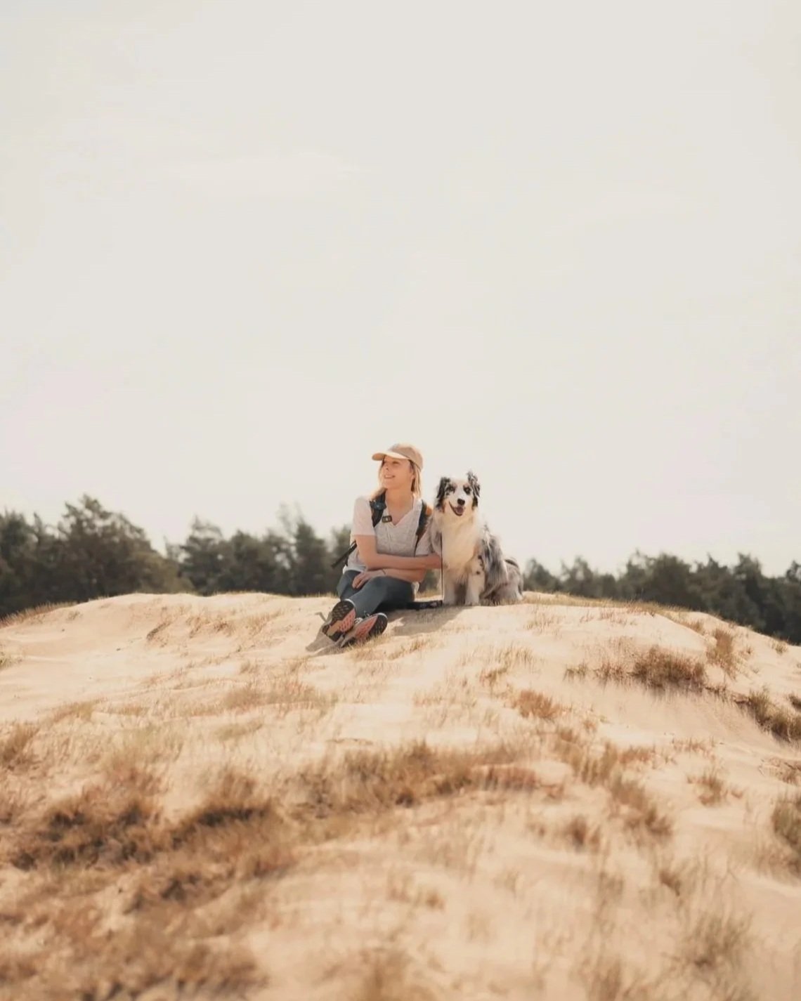Barbara sits on a sand dune on the Veluwe with her Australian Shepherd Ekko, in an open field with trees.