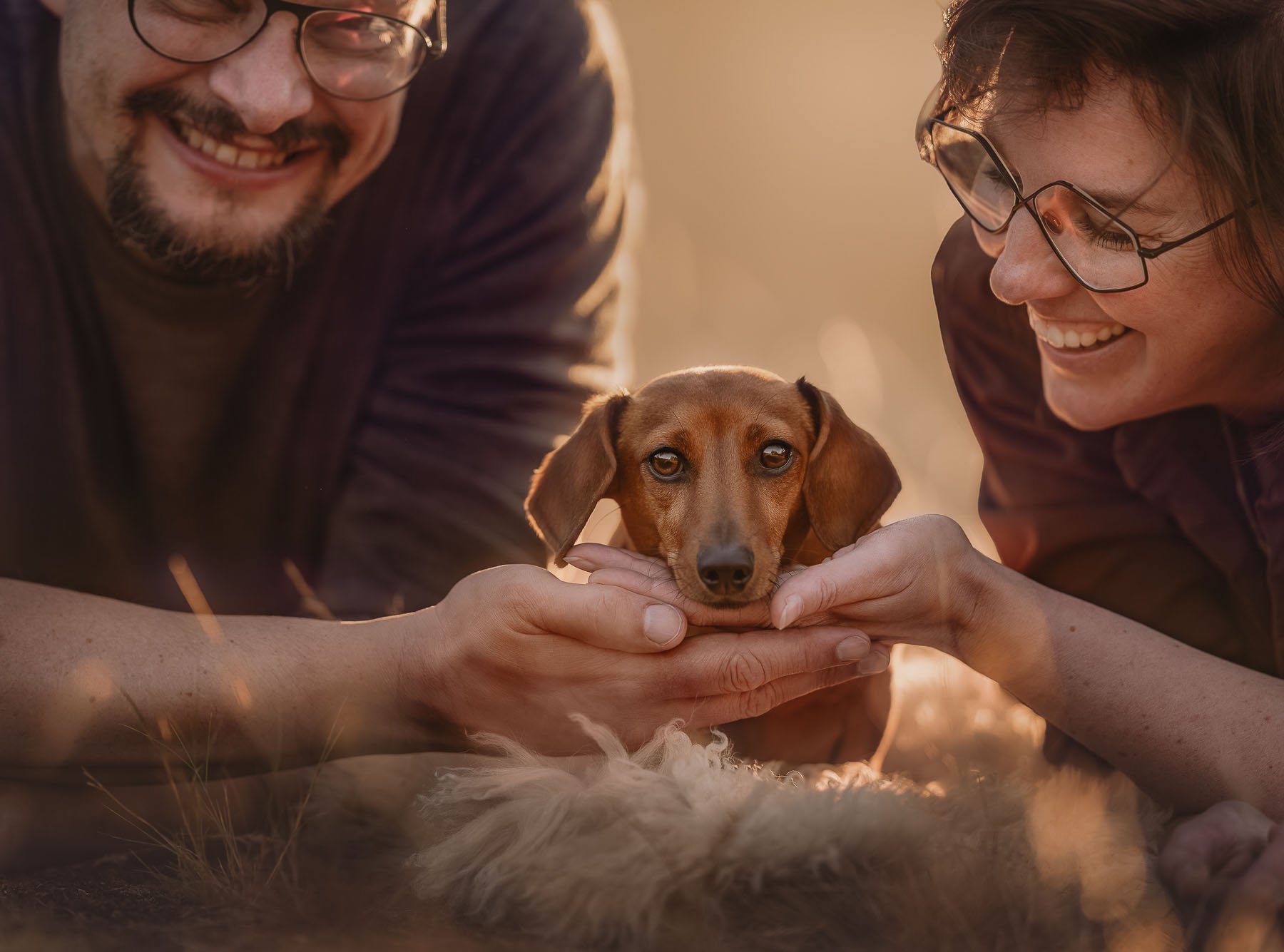 Two people smiling as they hold a brown dachshund, its head between their hands, outdoors at sunset.