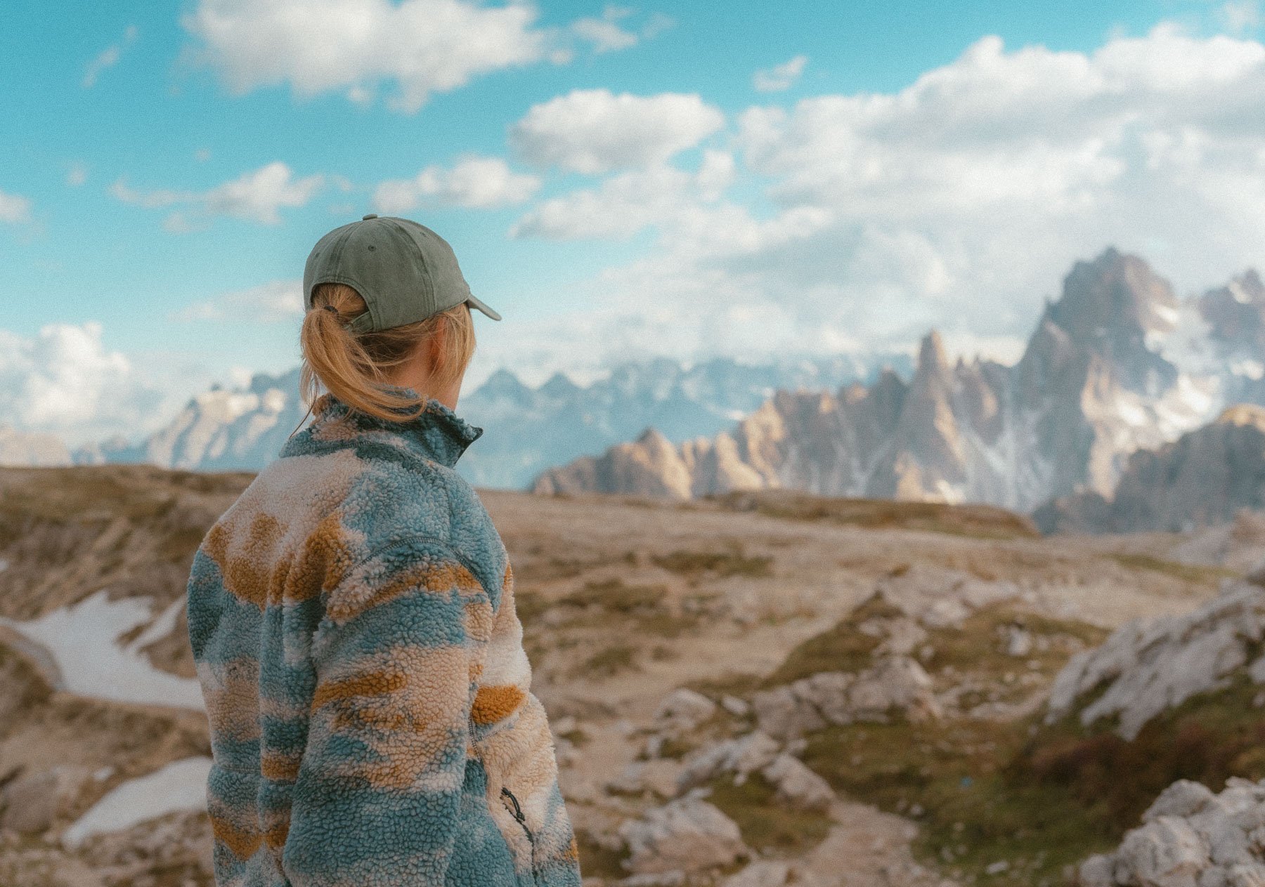 Person in a grey cap looks out over a mountainous landscape with blue skies and clouds.