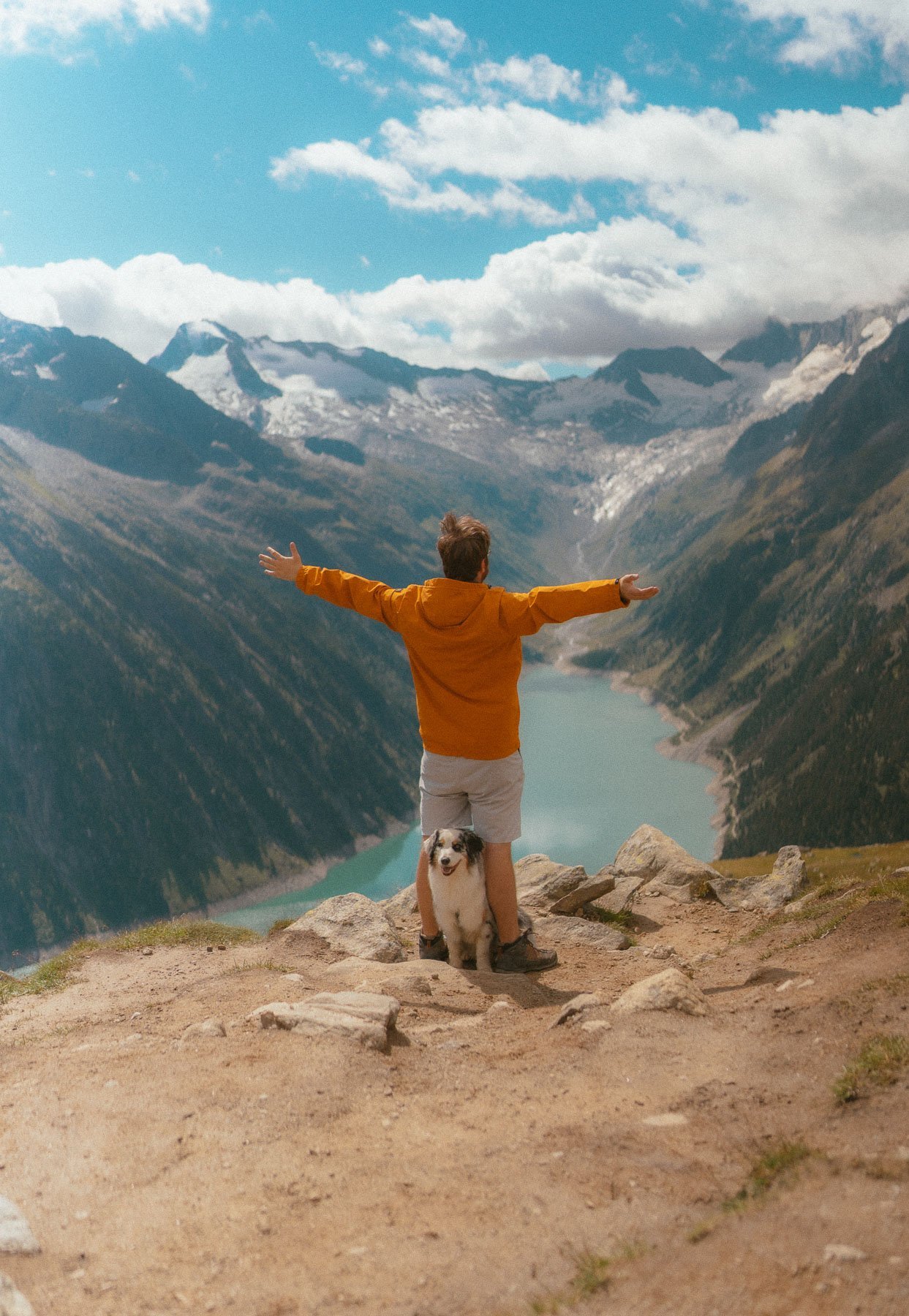 Man in an orange jacket with his arms spread wide, next to a dog, on a mountain overlooking a fjord and snow-capped peaks under a blue sky with clouds.